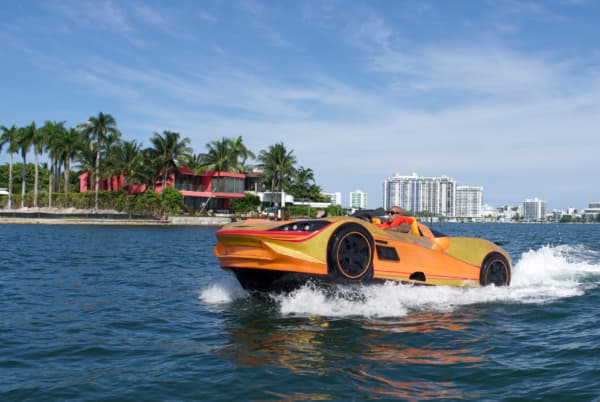 Orange car-shaped boat speeding across blue water past palm trees and a pink house.