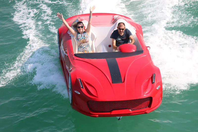 Red sports car-shaped boat speeding through turquoise water with a man driving and woman cheering.