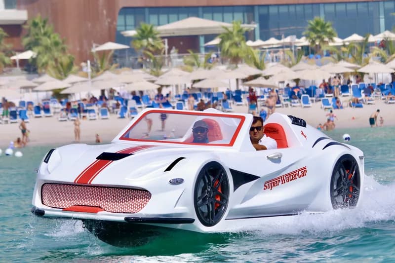 White sports car-shaped boat with red stripes cruising on water near a busy beach.